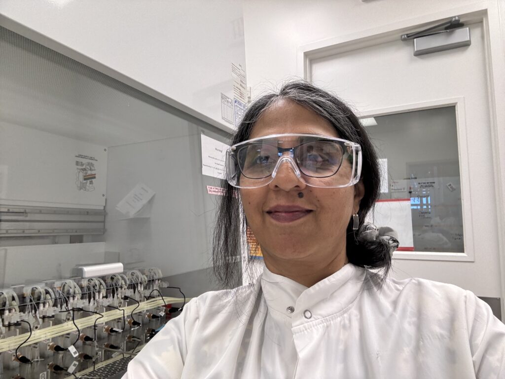 A selfie of Sheela Vemu in a white lab coat and safety glasses 