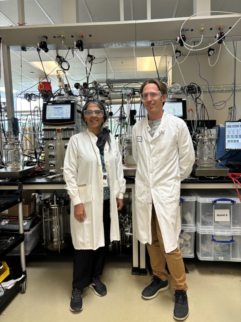 Two researchers in white lab coats stand in front of fermentation equipment in a laboratory